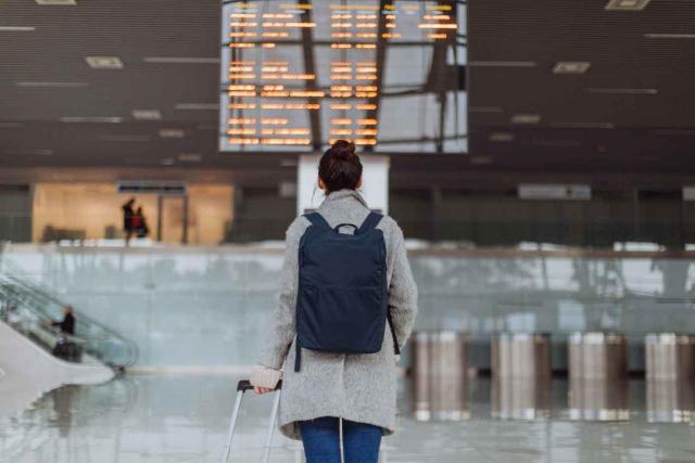 empresas de trabajo temporal; seguridad social; trabajadores cedidos. Chica de espaldas con una mochila y una maleta de carro, comprobando su vuelo en una pantalla