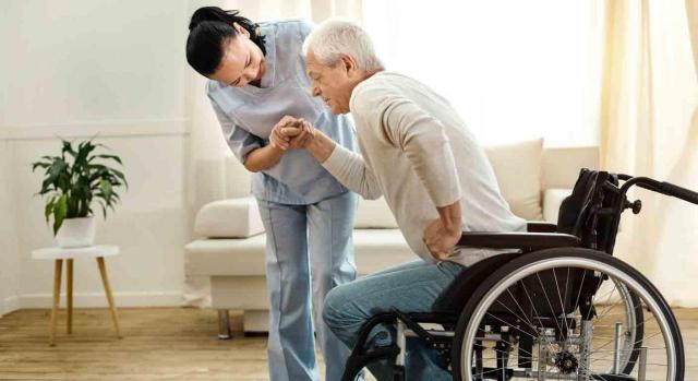 Gran invalidez. Sanitaria ayudando a un hombre mayor a levantarse de la silla de ruedas