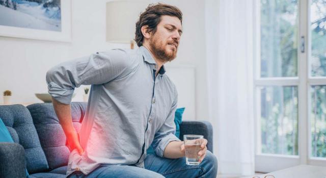 Perseguía el objetivo legítimo de proteger la productividad de la empresa y la eficiencia en el trabajo. Imagen de joven con dolor lumbar sentado en el sofá con un vaso de agua en la mano