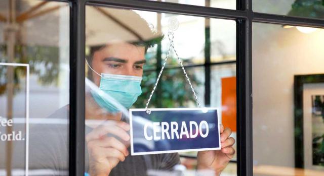 Despido objetivo en tiempos de COVID-19. Chico con mascarilla poniendo el cartel de Cerrado en la puerta de un negocio