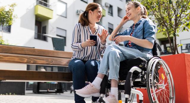 El empleo de una asistente personal que ayude a una persona con discapacidad en la vida cotidiana puede reservarse a las personas de la misma franja de edad. Imagen de dos chicas, una sentada en un banco de la calle y la otra en una silla de ruedas