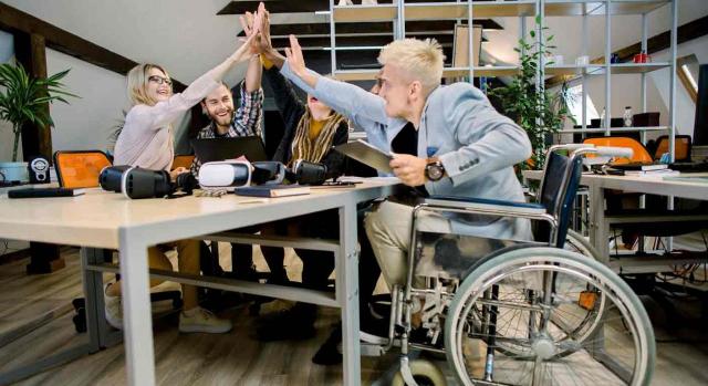 Personas con Discapacidad. 5 compañeros alrededor  de la mesa en la oficina, chocan las manos sonrientes, uno de ellos sentado en una silla de ruedas