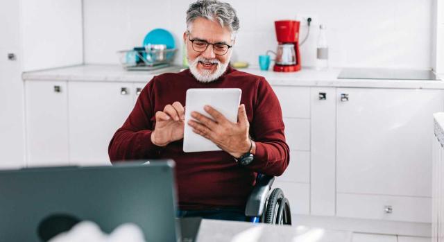 Acceso al subsidio para mayores de 52 años por beneficiario de una previa pensión de incapacidad permanente total (IPT): para cubrir el requisito de 15 años de carencia pueden computarse las cotizaciones anteriores a la IPT. Imagen de un hombre en sillas de ruedas revisndo su correo electrónico en su cocina