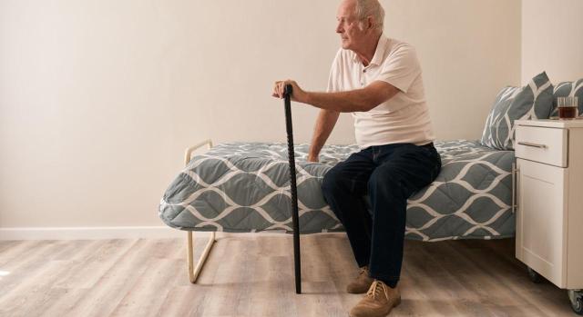 Pensiones no contributivas. El arriendo de una habitación en un piso compartido también da derecho al complemento para el alquiler de vivienda. Imagen de un abuelo con bastón sentado en una cama