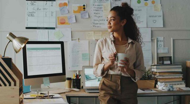 Una mujer toma un café sonriente de espaldas a su mesa de trabajo