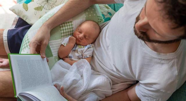 La Audiencia Nacional establece que las empresas no tienen que retribuir el permiso parental de 8 semanas. Imagen de un papa leyendo un libro mientras tiene a su bebe en su regazo