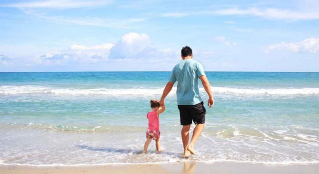 Planificación vacaciones en la empresa. Padre con su pequeña de la mano andando por la orilla del mar