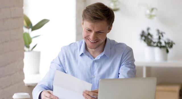 Pagas extraordinarias. Hombre sonriente con un papel en la mano sentado frente a un escritorio