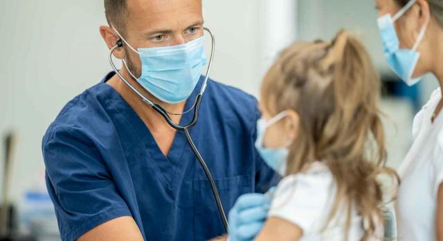 Comunidad de Madrid; médicos de familia; prevención de riesgos laborales. Médico pediatra examinando a una niña sentada en el regazo de su madre y todos con mascarillas