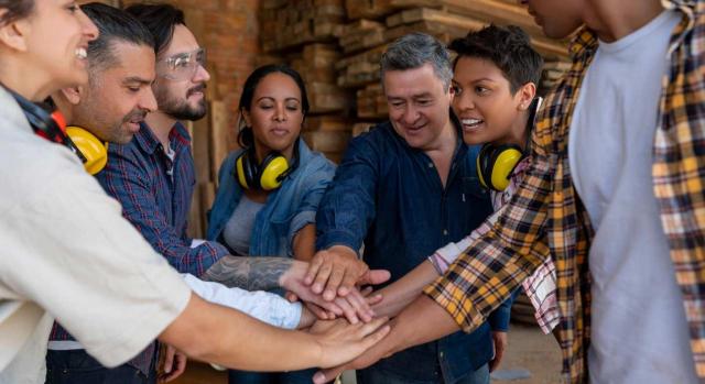 La negociación de los planes de igualdad se rige por las previsiones referidas a la negociación colectiva. Imagen de grupo de trabajadores en una fábrica poniendo sus manos juntas en concepto de Equipo