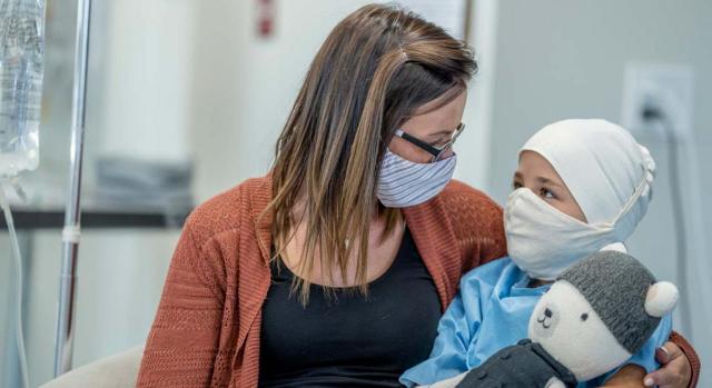 El acceso a la prestación está condicionado a que ambos progenitores trabajen, sin excepciones. Imagen de madre con bebé en brazos en un entorno hospitalario y ambos con mascarilla