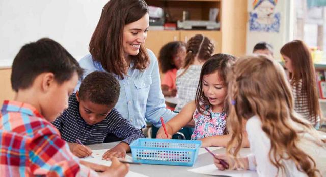 Modificación sustancial de las condiciones de trabajo; horario; impacto de género; conciliación de la vida laboral y familiar. Una profesora observa sonriente como trabajan en una mesa redonda sus alumnos pequeños