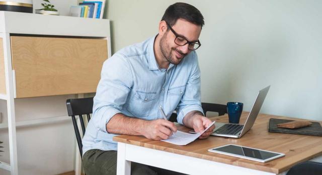 Reducciones y bonificaciones en las cuotas a la Seguridad Social de los autónomos. Imagen de un hombre sonriente en una mesa de escritorio apuntando algo en un papel y su tablet y portátil al lado