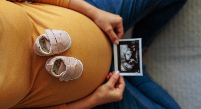 Adhesión de España al Convenio OIT 183 (2000) sobre protección de la maternidad. Imagen de unos zapatitos sobre la barriga de una mujer embarazada mientras ve la foto de una ecografía